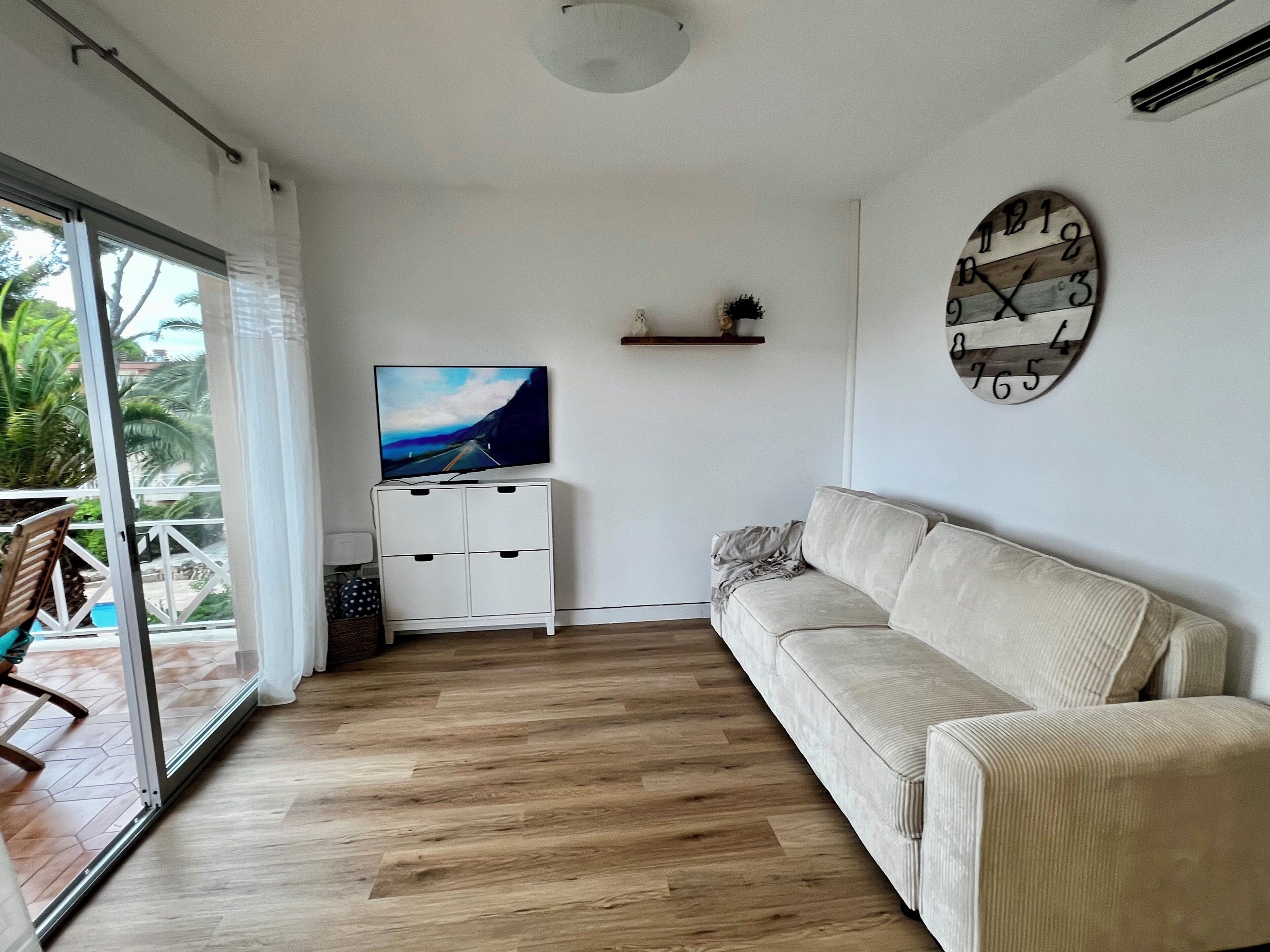 A living room with a sofa, a TV on a cabinet, a clock on the wall, and a view through a glass door.