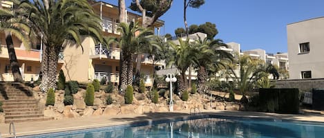 A swimming pool with clear blue water, surrounded by palm trees and a building with balconies.