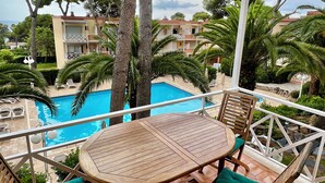 Une table en bois, agrémentée de coussins verts, est placée sur un balcon donnant sur une piscine, entourée de palmiers et avec un bâtiment visible au loin.