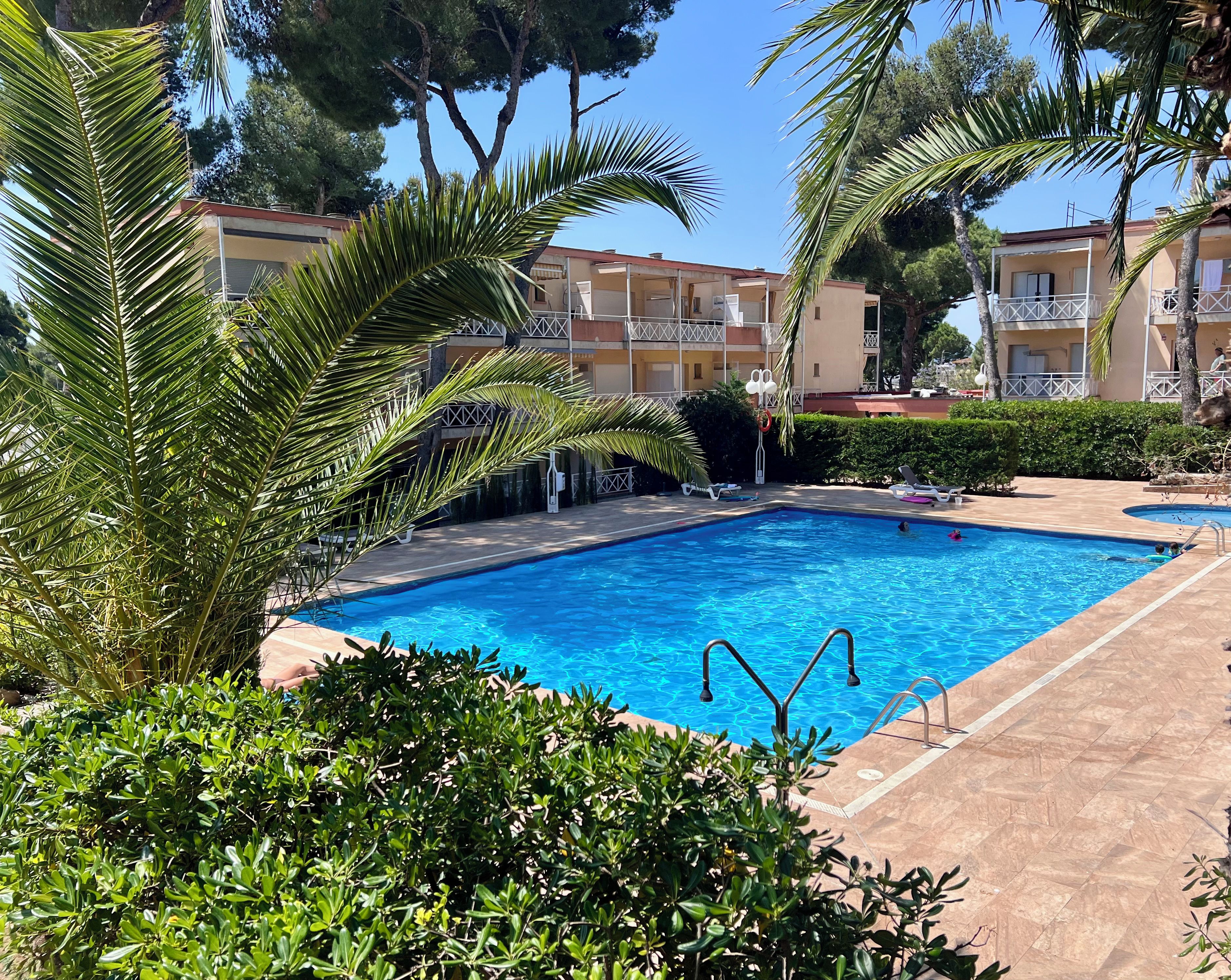 A swimming pool with clear blue water, surrounded by lush greenery and a multi-story building with balconies.