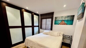 Une chambre à coucher avec un lit, un plancher en bois, une fenêtre avec des stores, un tableau au mur et une petite table avec une chaise.