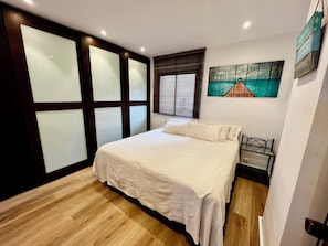 A bedroom with a bed, wooden flooring, a window with blinds, a painting on the wall, and a small table with a chair.
