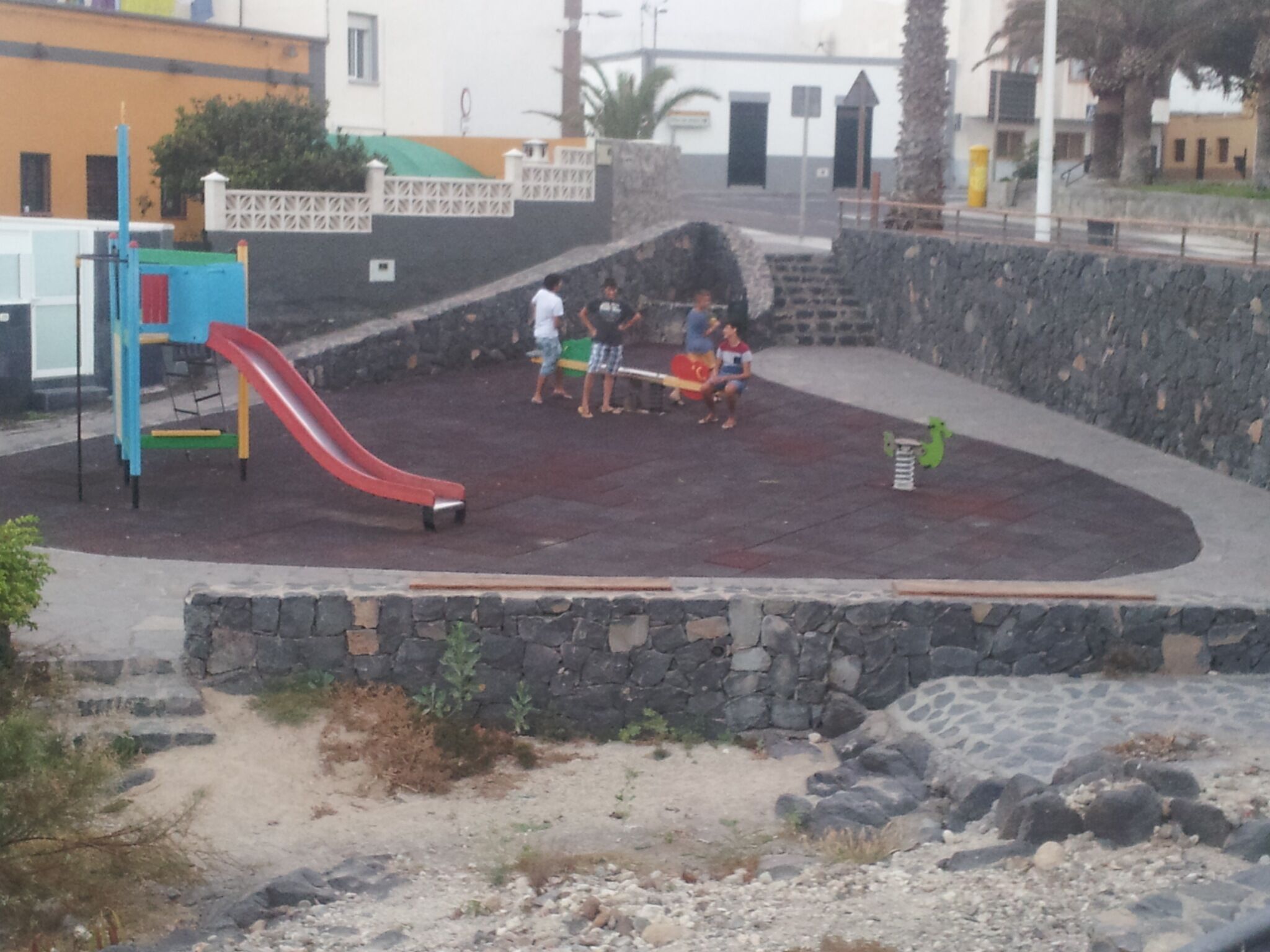 Une aire de jeux avec un toboggan rouge et une structure d’escalade, entourée d’un mur de pierre et d’une zone pavée.
