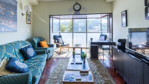 Ein Wohnzimmer mit einer Couch, einem Couchtisch und einem Fernseher. Ein Fenster bietet Blick auf eine Stadtlandschaft.