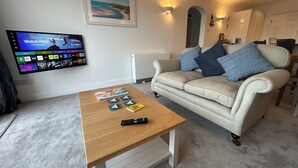 A living room with a flat-screen TV displaying a streaming service interface, a beige sofa with blue cushions, a wooden coffee table with remote controls, and a framed picture on the wall. - Luxury Apartment, Magnificent Sea Views, Private Access to Beach, Bigbury-on-Sea (Bigbury on Sea)