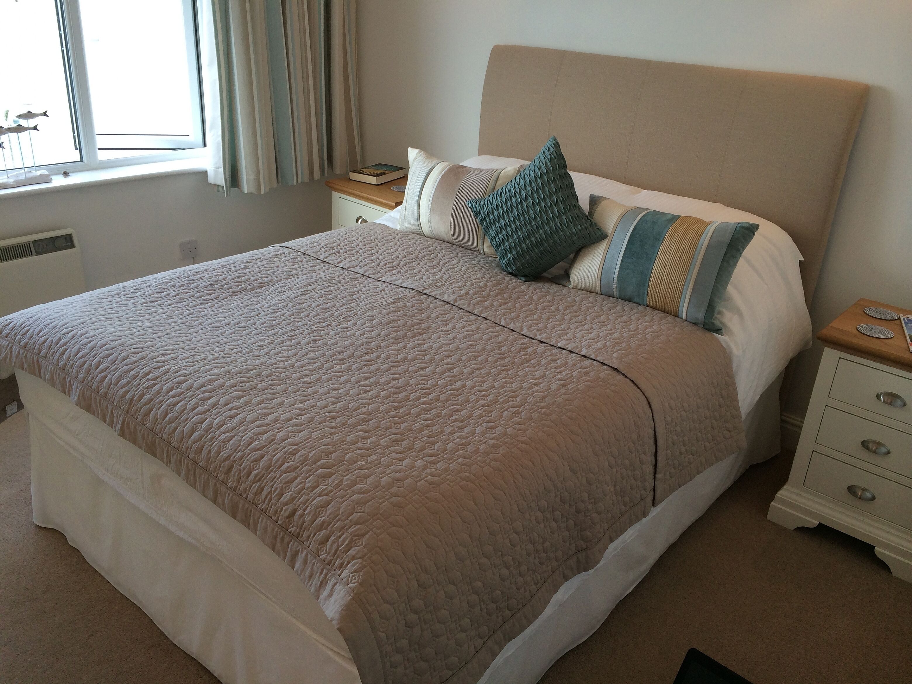 Een net opgemaakt bed met een beige deken, een kussen met een geometrisch patroon en een nachtkastje met een boek en een decoratief object.