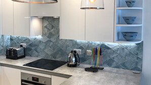 A modern kitchen with white cabinets, a black stove, and a coffee maker. There are two pendant lights with exposed bulbs. - Luxury Apartment, Magnificent Sea Views, Private Access to Beach, Bigbury-on-Sea (Bigbury on Sea)