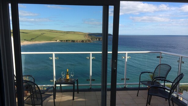 A balcony with a table, two chairs, and a bottle of wine, overlooking a coastal landscape with the sea and hills. - Luxury Apartment, Magnificent Sea Views, Private Access to Beach, Bigbury-on-Sea (Bigbury on Sea)