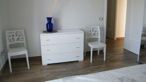 Un dormitorio con cama, cómoda blanca, dos sillas y un jarrón azul.