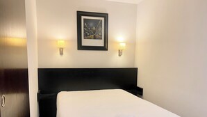 Hotellihuone, jossa on siististi pedattu sänky, kaksi seinälle asennettua lamppua ja sängyn yläpuolella oleva kehystetty kuva.