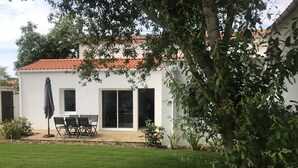 Un bâtiment blanc avec un toit en tuiles, une terrasse avec une table et des chaises, et une cour gazonnée.