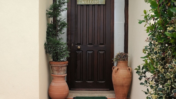 A dark wooden door with a 'WELCOME' sign above it, flanked by two large terracotta pots and a small green mat on the tiled patio. - Luxury Villa with Private Pool, Panoramic Views, Solarium Terrace, BBQ, Parking (Pisa)