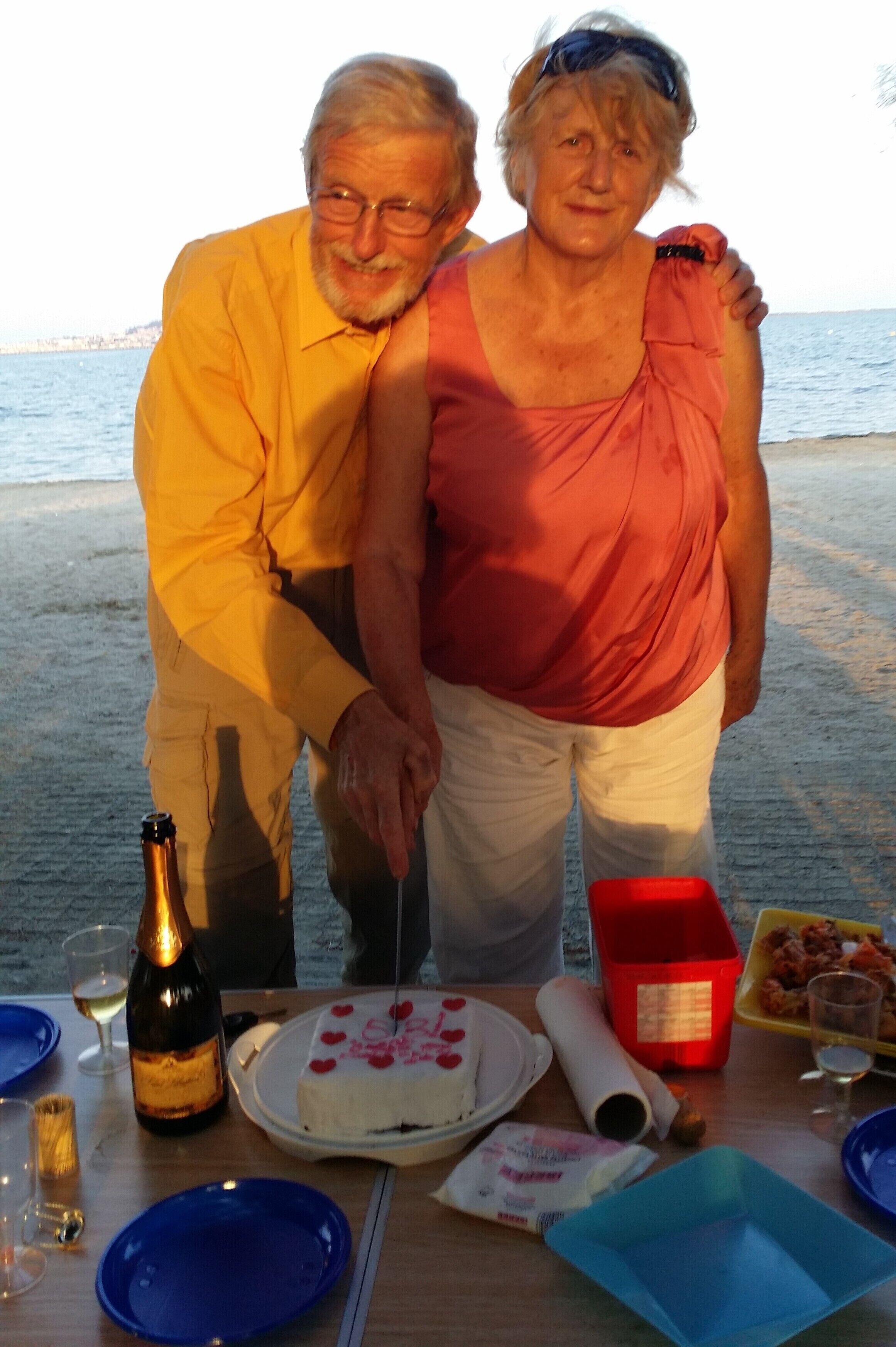 A dining table with a bottle, glasses, and a cake on a beach setting.