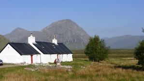 A white building with a black roof and red door, surrounded by grass and mountains.