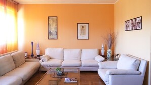 A living room with a beige sofa, a glass coffee table, and a white armchair. There are framed artworks on the wall and a vase with branches on a side table.