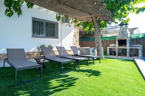 Un patio trasero con cuatro sillones de jardín, una zona de barbacoa de piedra y una piscina pequeña.