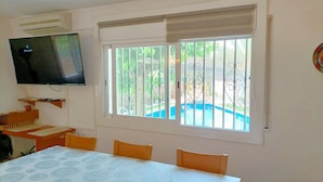 Ein Zimmer mit Esstisch, Stühlen, einem an der Wand montierten Fernseher und einem Fenster mit Blick auf einen Swimmingpool.