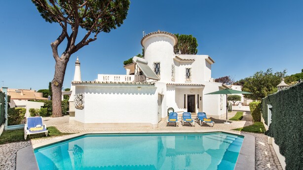A white building with a pool, blue lounge chairs, and a tree.