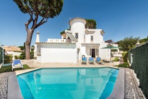 Un bâtiment blanc avec une piscine, des chaises longues bleues et un arbre.