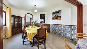A dining area with a table set for four, wooden chairs, a yellow tablecloth, a vase with flowers, and a painting on the wall.
