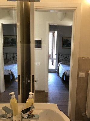 Ein Badezimmer mit einem großen Spiegel, einem Waschbecken mit zwei Seifenspender und zwei Betten, die durch eine offene Tür sichtbar sind.