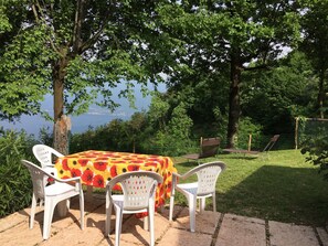Ein Tisch mit einer blumigen Tischdecke und weißen Plastikstühlen, auf einer Steinterrasse mit Blick auf ein Gewässer und Bäume im Hintergrund.
