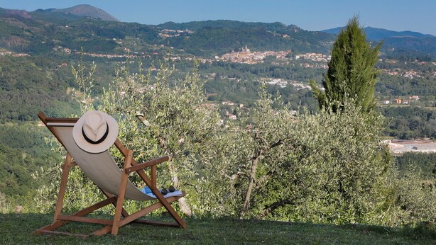 Ein Holzstuhl mit Hut und Sonnenbrille, draußen platziert mit Blick auf eine Stadt und Hügel im Hintergrund.