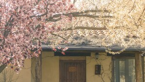 Un edificio con puerta de madera, dos sillas, una mesa y una pérgola con árboles en flor.