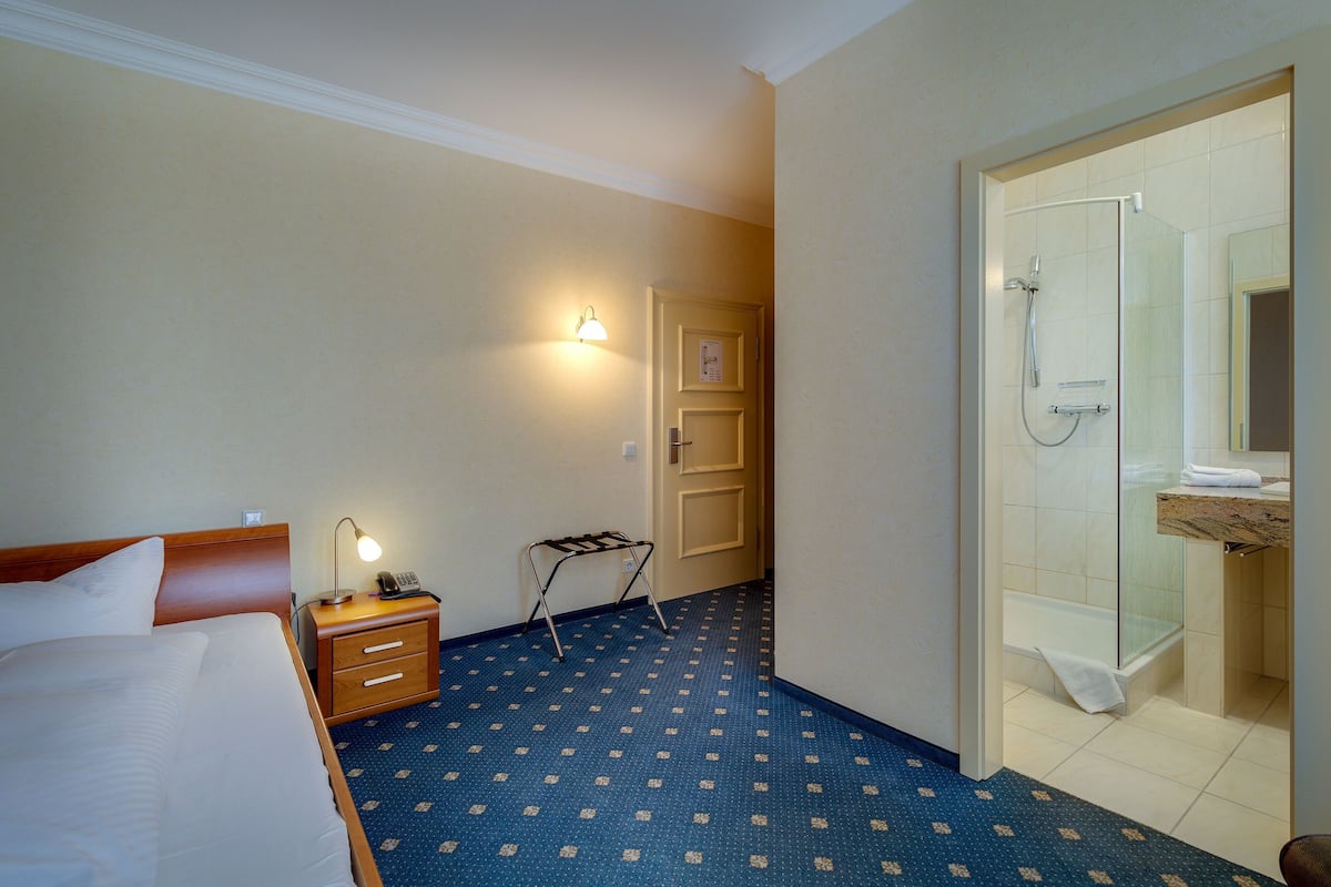 A hotel room with a bed, bedside lamp, a small table with a lamp, a wall-mounted light, a shower area with a glass door, and a bathroom with a sink and mirror.