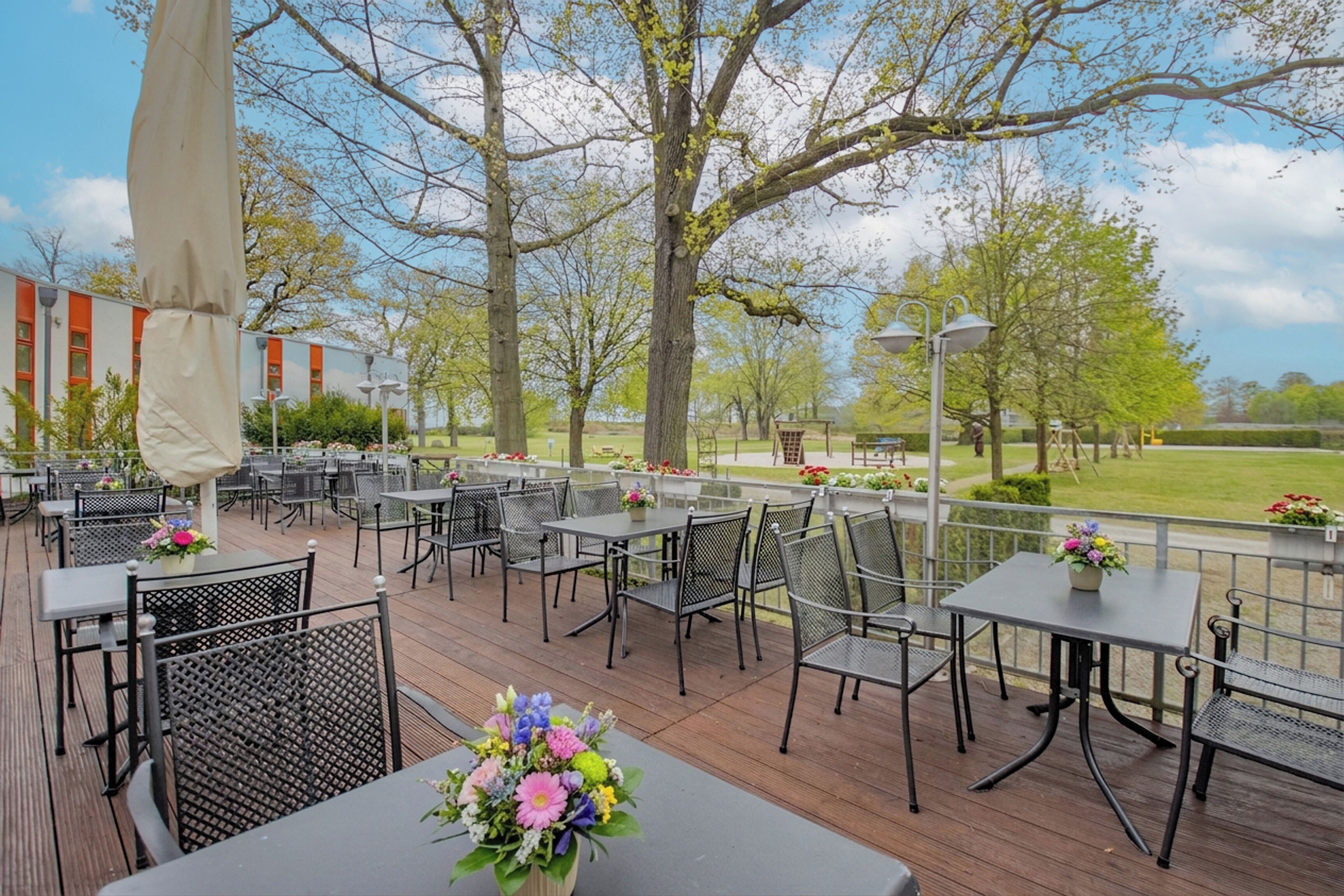 Une terrasse extérieure avec des tables et des chaises, entourée d’arbres et bordée d’un bâtiment à la façade rouge et blanche.
