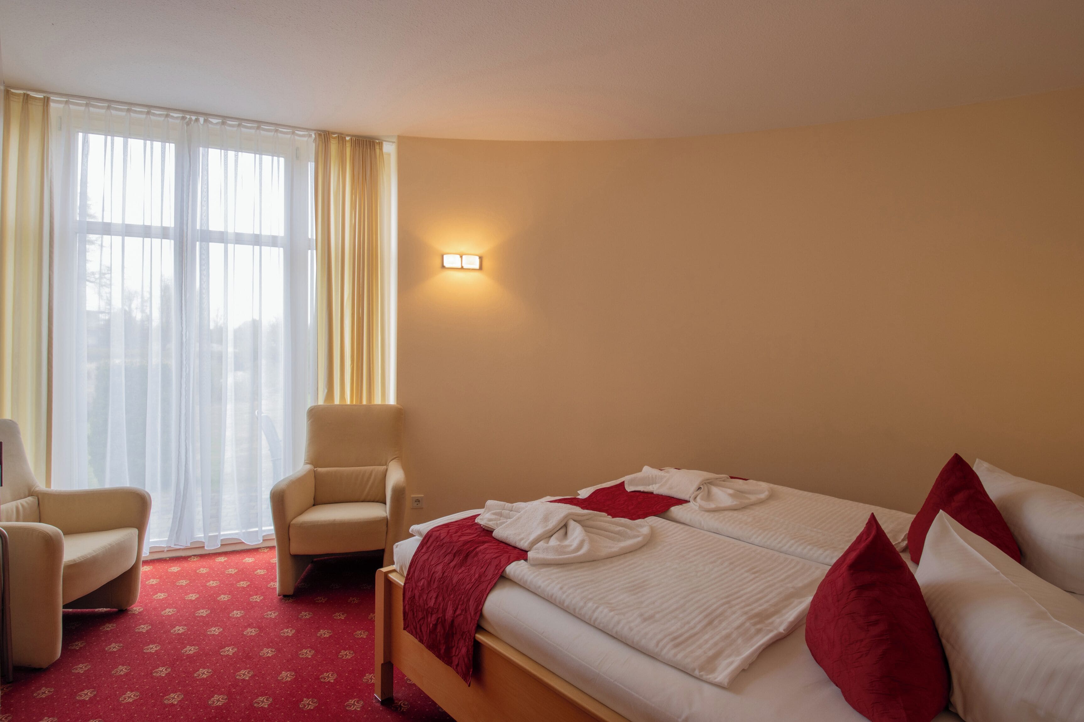 Una habitación de hotel con una cama, dos sillas, un ventanal con cortinas y una lámpara fijada en la pared.