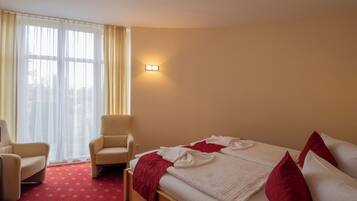 Una habitación de hotel con una cama, dos sillas, un ventanal con cortinas y una lámpara fijada en la pared.