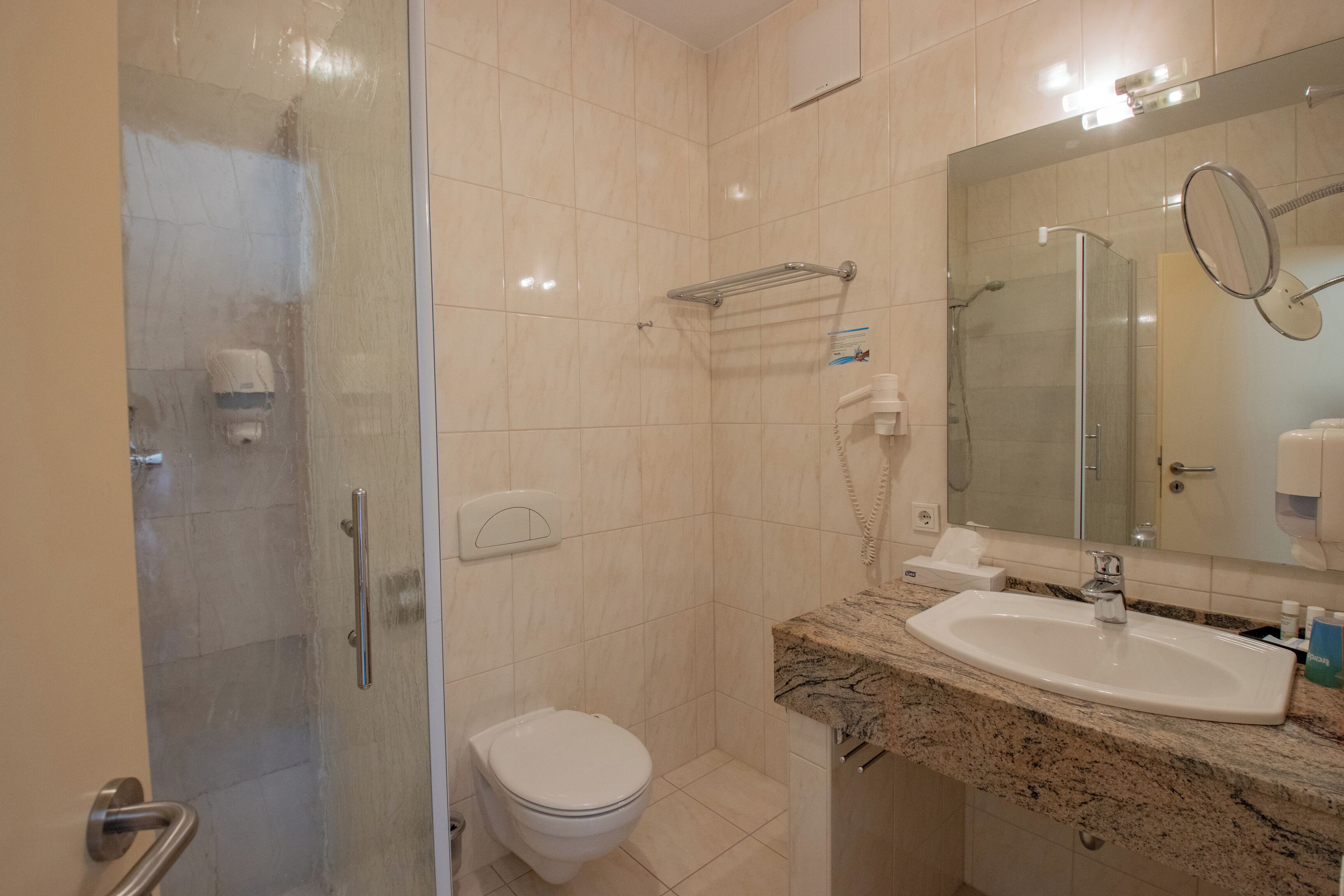 Un baño con ducha de vidrio, inodoro blanco, lavamanos con encimera de mármol y espejo con marco redondo.