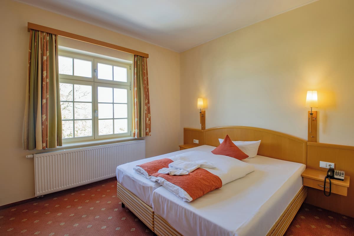 A hotel room with a double bed, a window with curtains, a bedside table with a lamp, and a radiator.