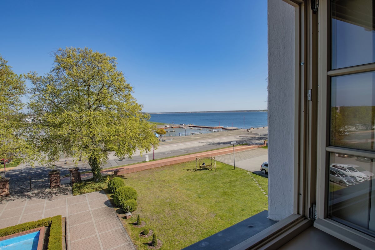 A view from a window overlooking a grassy area with a tree, a pool, and a body of water.
