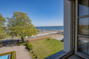 A view from a window overlooking a grassy area with a tree, a pool, and a body of water.