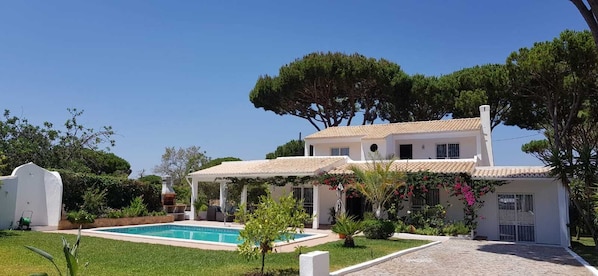 Una casa de dos plantas con piscina, rodeada de follaje y una pérgola.