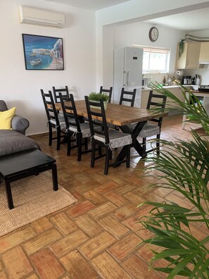 A dining area with a wooden table and chairs, a grey sofa, a rug, and a potted plant.