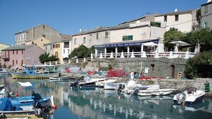 Un port de plaisance où sont amarrés différents types de bateaux, un restaurant doté d’une terrasse, et des bâtiments avec des balcons donnant sur l’eau.