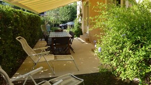 Une terrasse extérieure avec une table et des chaises, un auvent jaune et de la verdure tout autour.
