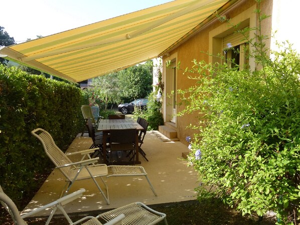 Eine Terrasse im Freien mit Tisch und Stühlen, einem gelben Baldachin und umgebender Bepflanzung.