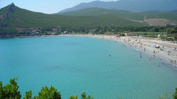 Une plage côtière aux eaux turquoise et limpides, bordée d’un rivage sablonneux, avec des collines verdoyantes en arrière-plan.