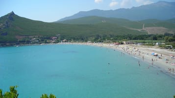 Une plage côtière aux eaux turquoise et limpides, bordée d’un rivage sablonneux, avec des collines verdoyantes en arrière-plan.