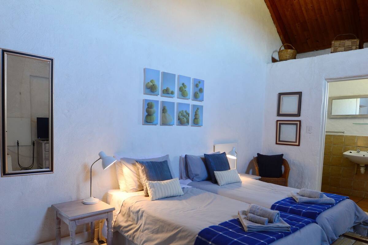 Ein Schlafzimmer mit zwei Betten, einer Holzbalkendecke, einer weißen Wand, einem Spiegel und einem kleinen Tisch mit einer Lampe.