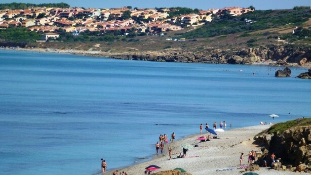 A coastal beach with people sunbathing and swimming, a rocky shoreline, and a town in the background.