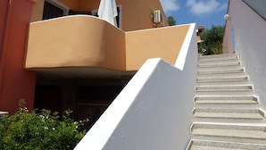 A staircase leading to a balcony with a white umbrella, surrounded by greenery and a clear blue sky.