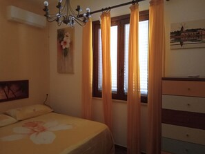 A bedroom with a bed, a dresser, a window with curtains, a chandelier, and a painting on the wall.