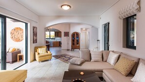 Uma sala de estar ampla com sofá, poltrona e mesa de centro. Uma cozinha é visível ao fundo, e há uma grande janela que traz luz natural.