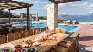 Uma área de refeições à beira da piscina, com mesa posta para uma refeição, piscina e vista para o mar.