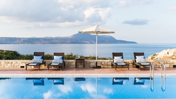 Uma área à beira da piscina com espreguiçadeiras e um guarda-sol, com vista para um corpo de água e terras distantes.